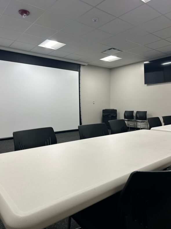 Duemelands Commercial Real Estate Celly Room B with tables and chairs set up and a projector screen hanging down from the ceiling 